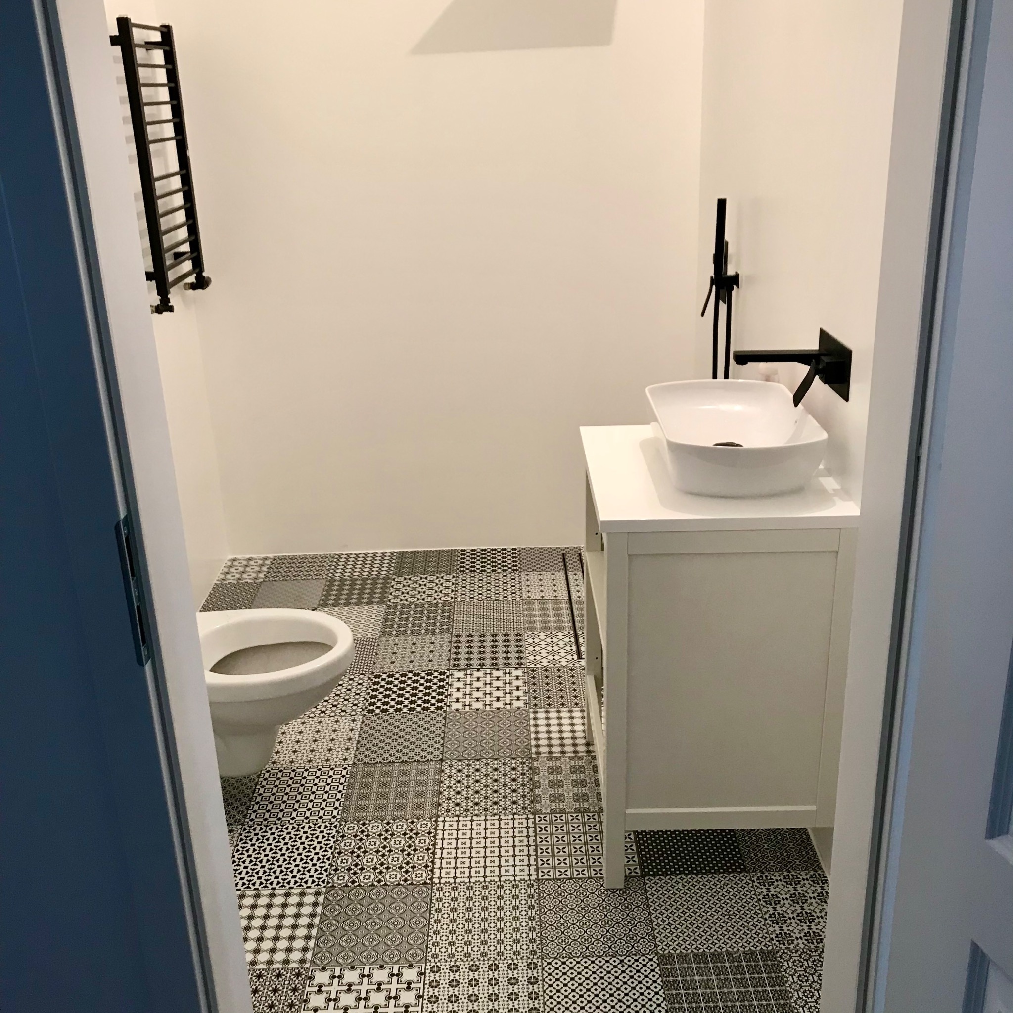 Nowoczesna łazienka z białą umywalką nablatową, czarną armaturą i grzejnikiem drabinkowym, wyróżniająca się patchworkową posadzką z płytek w odcieniach szarości i czerni.