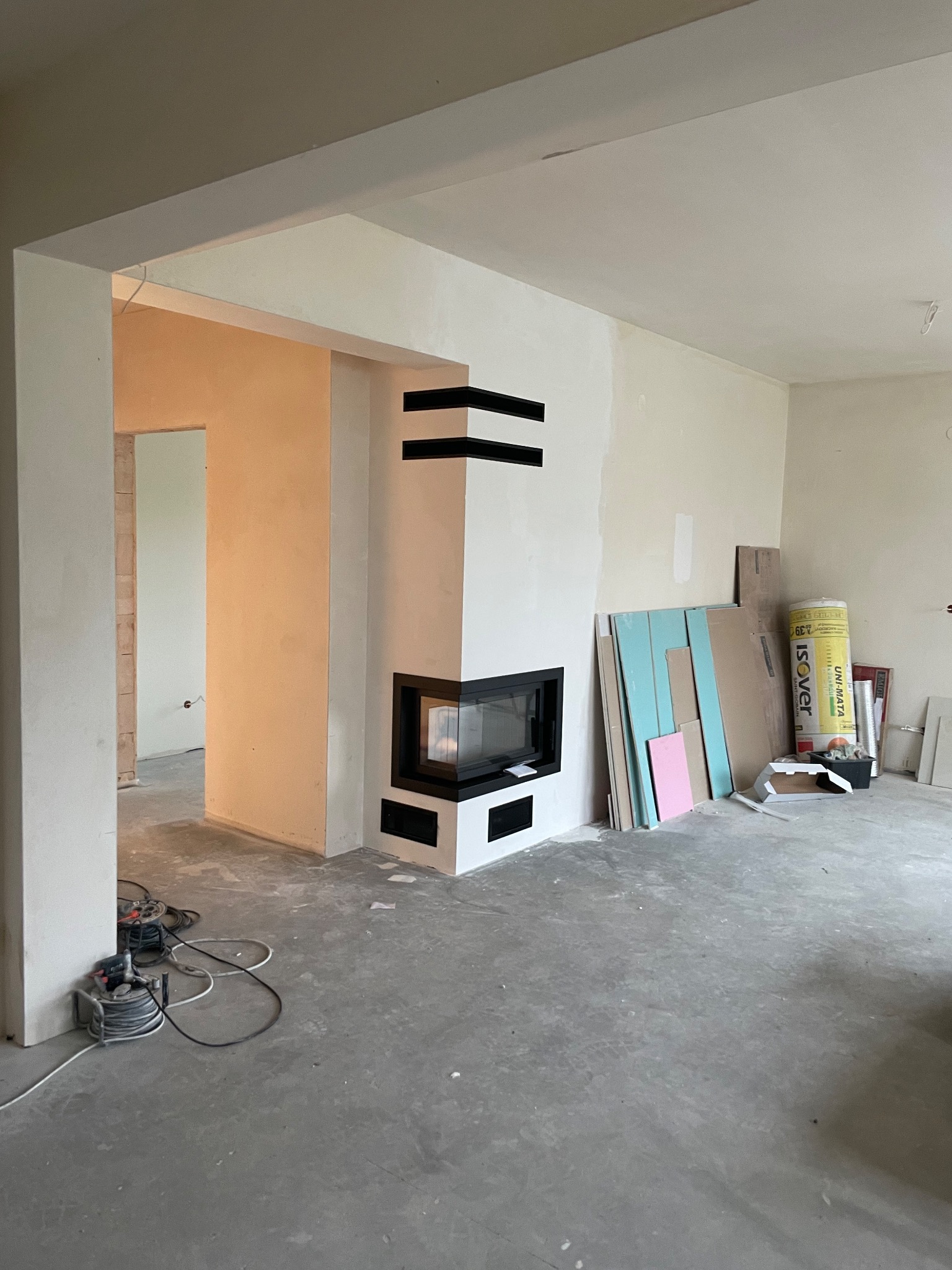 Wnętrze w trakcie remontu z narożnym kominkiem o czarnych, poziomych dekoracjach, widoczne płyty gipsowe i rolka izolacji przy ścianie, surowy beton na podłodze.