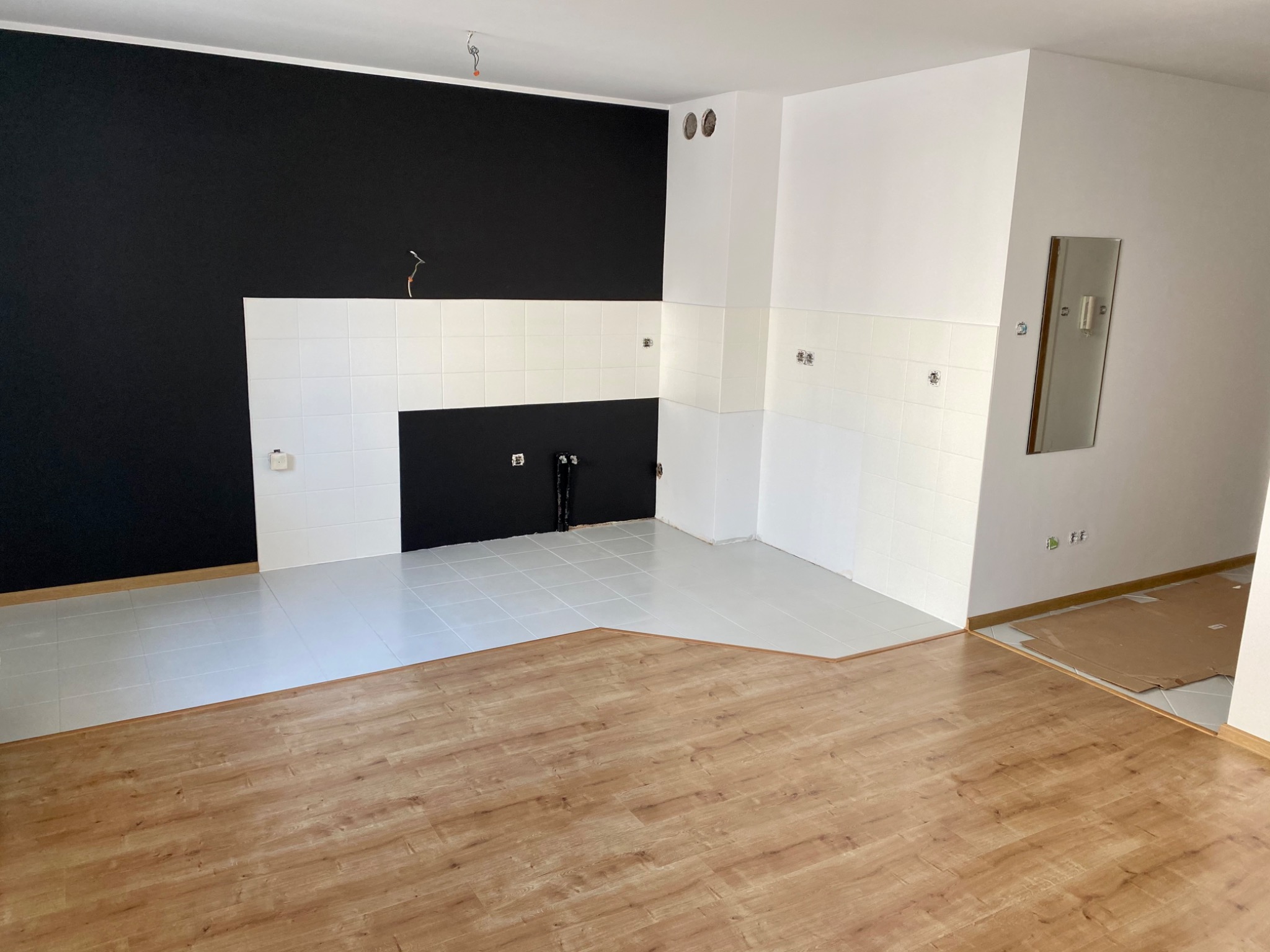 Wnętrze w trakcie prac: kontrastowa ściana pomalowana na czarno, białe płytki na ścianie i podłodze, fragment ułożonych paneli podłogowych w odcieniach brązu.