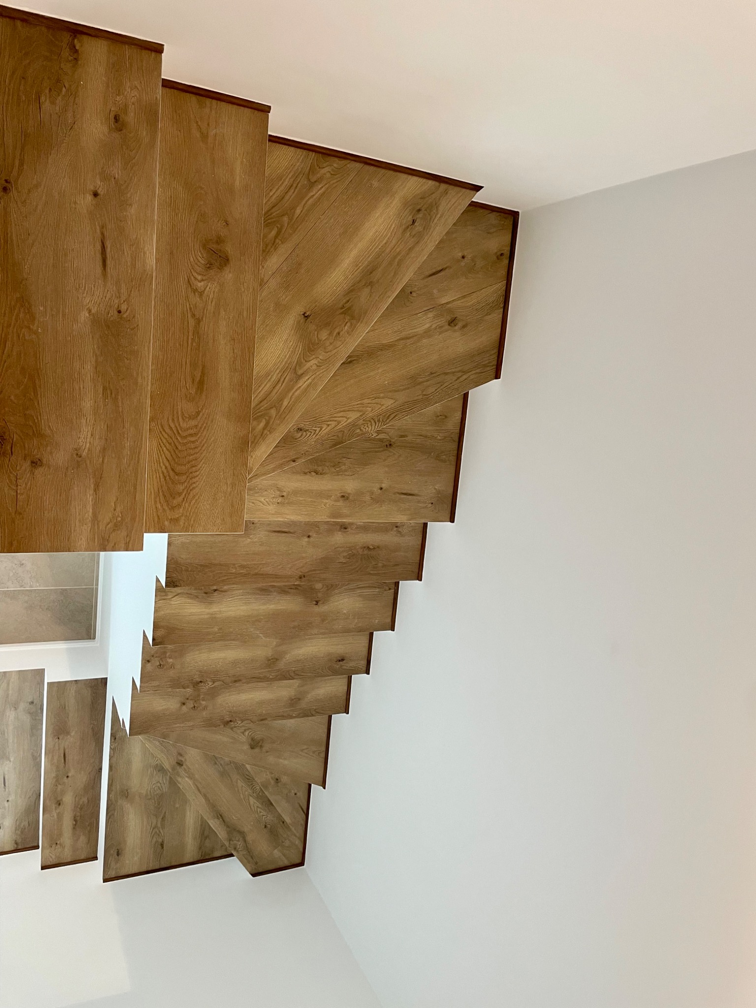 Widok z góry na drewniane schody spiralne z brązowymi krawędziami, kontrastujące z białymi ścianami i sufitem, ukazujące precyzję montażu i estetyczne wykończenie.