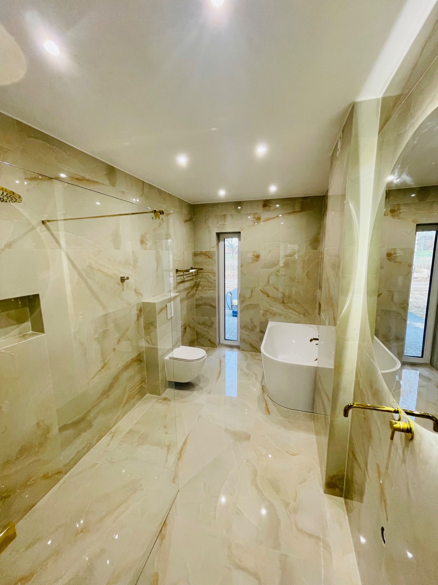 Nowoczesna łazienka z marmurowymi płytkami w odcieniach beżu i złota, z wolnostojącą wanną, toaletą podwieszaną i szklaną kabiną prysznicową typu walk-in, oświetlona punktowymi lampami sufitowymi.