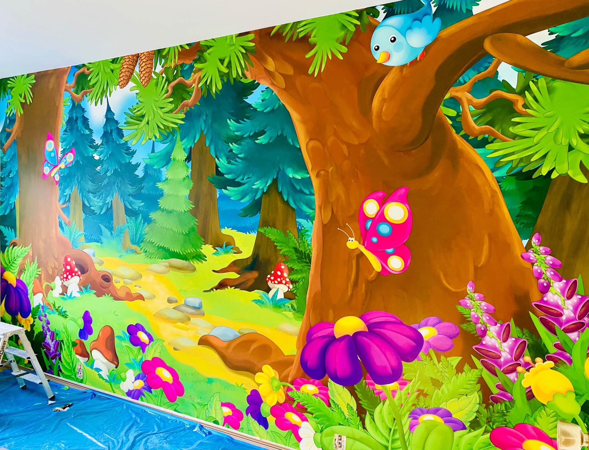 Barwny, ręcznie malowany mural na ścianie przedstawiający leśny krajobraz z drzewami, kwiatami, grzybami, motylami i ptakiem, zabezpieczony folią i z rozłożoną drabiną w rogu pokoju.