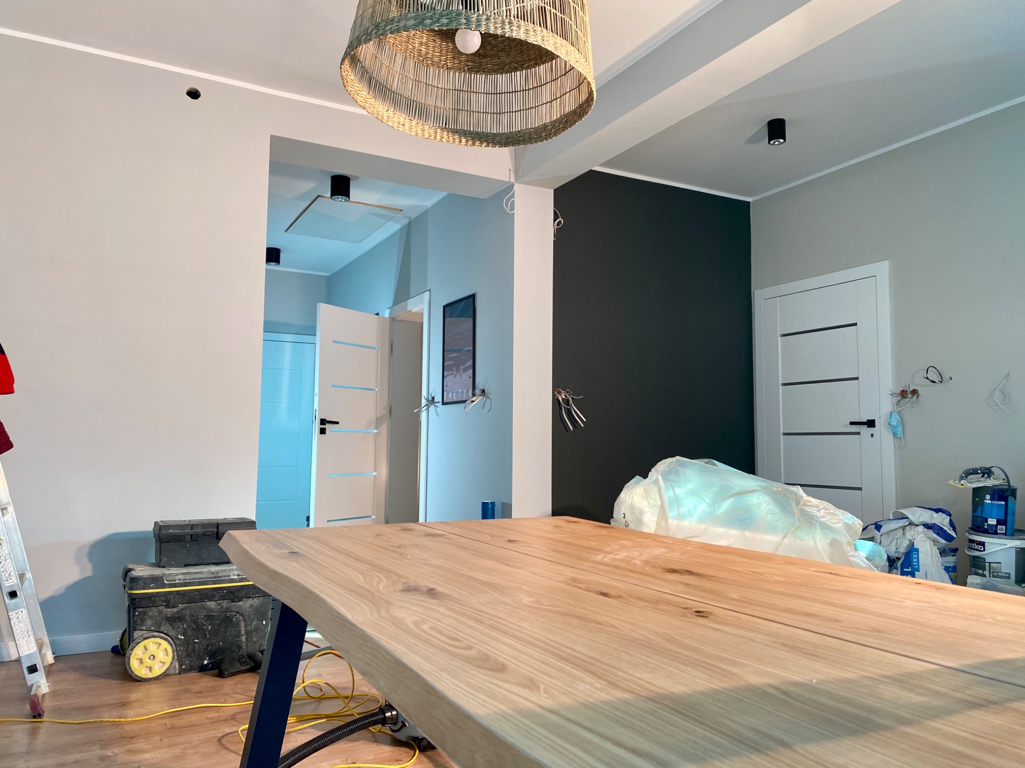 Wnętrze w trakcie odświeżania: widoczna duża drewniana płyta stołu na pierwszym planie, w tle drabina, skrzynki narzędziowe, pomalowana na ciemny kolor ściana, wiszące przewody elektryczne...