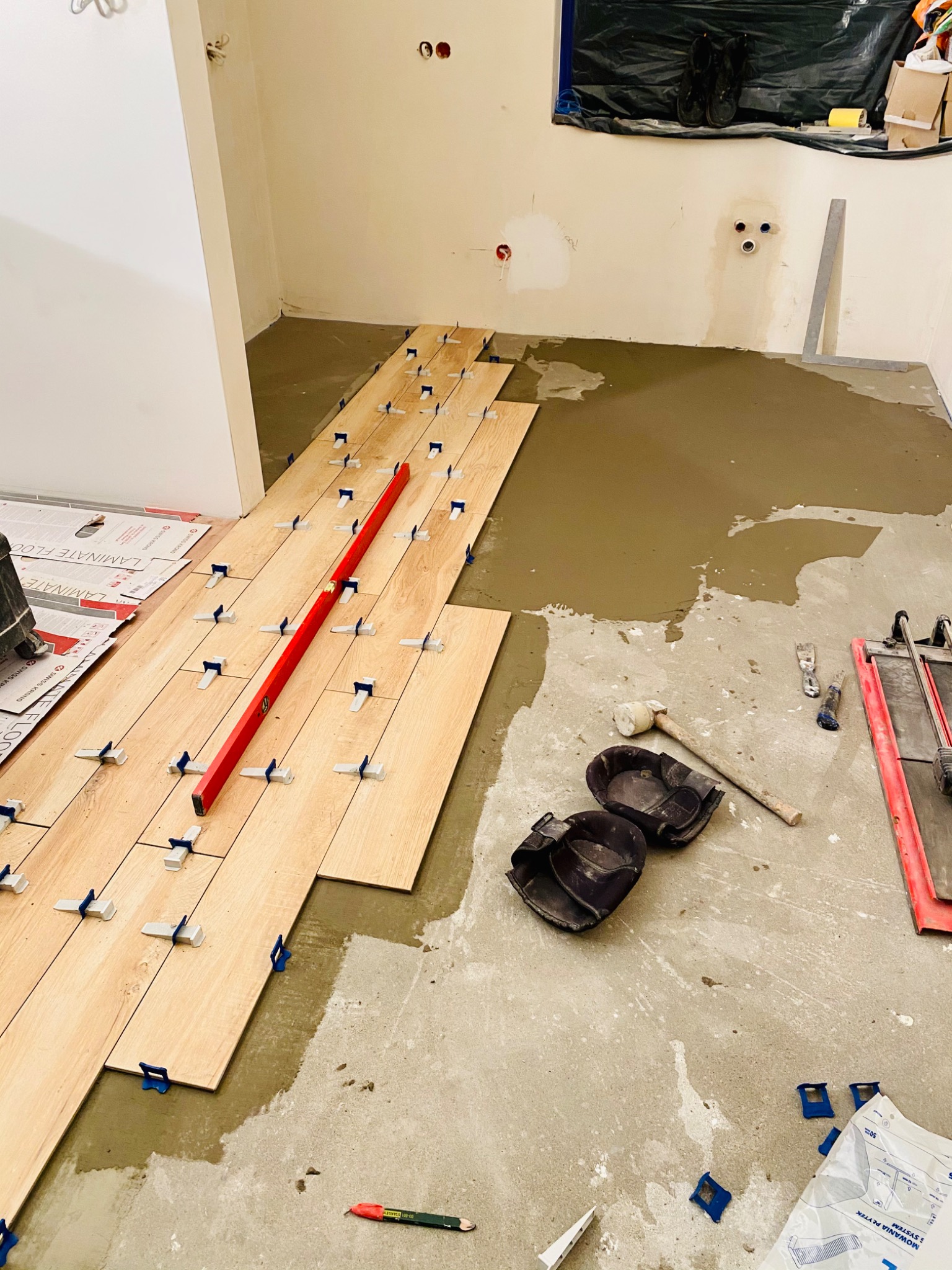 Ułożone rzędy drewnopodobnych płytek podłogowych z niebieskimi klipsami poziomującymi, czerwona poziomica, robocze nakolanniki i narzędzia na podłożu z wylanym podkładem, przygotowywane do dalszych...