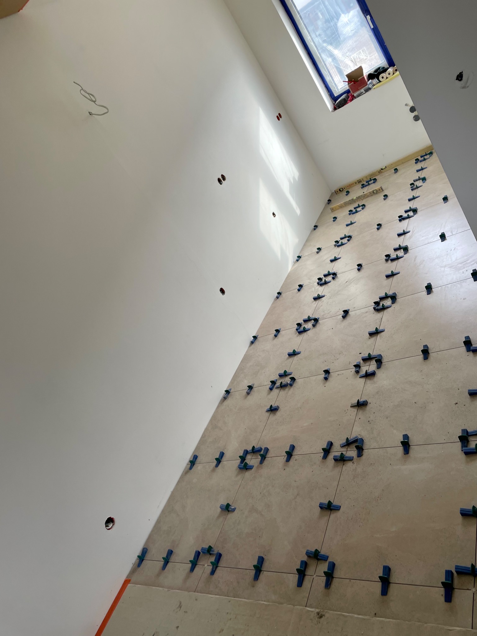 Ściana w trakcie układania płytek, z widocznymi niebieskimi klipsami poziomującymi, poziomica i okno dachowe w tle.
