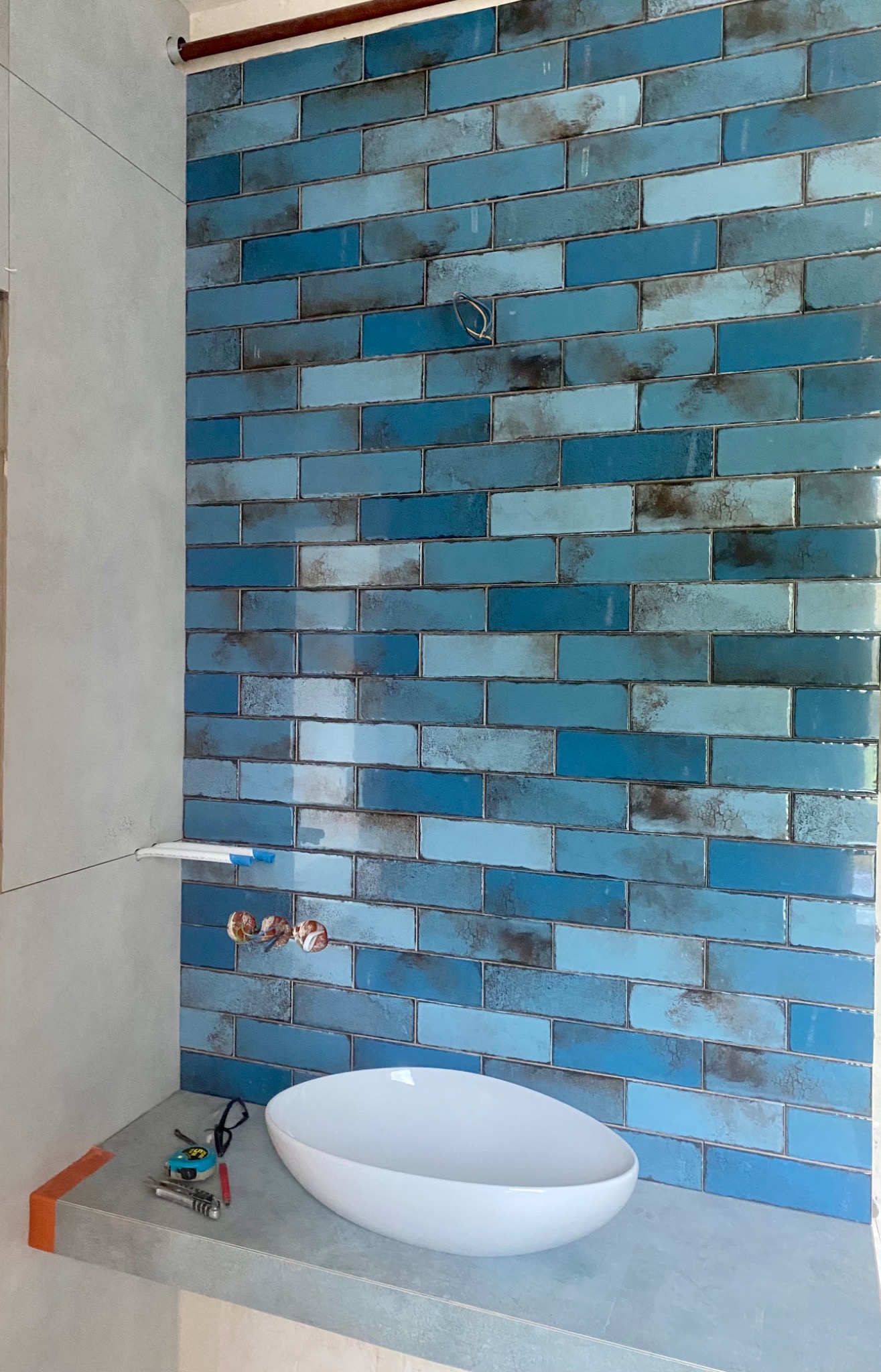 Ściana wyłożona błękitną, cegiełkową glazurą w łazience, z widocznymi wystającymi kablami i niedokończonym blatem pod umywalkę, na którym leżą narzędzia.