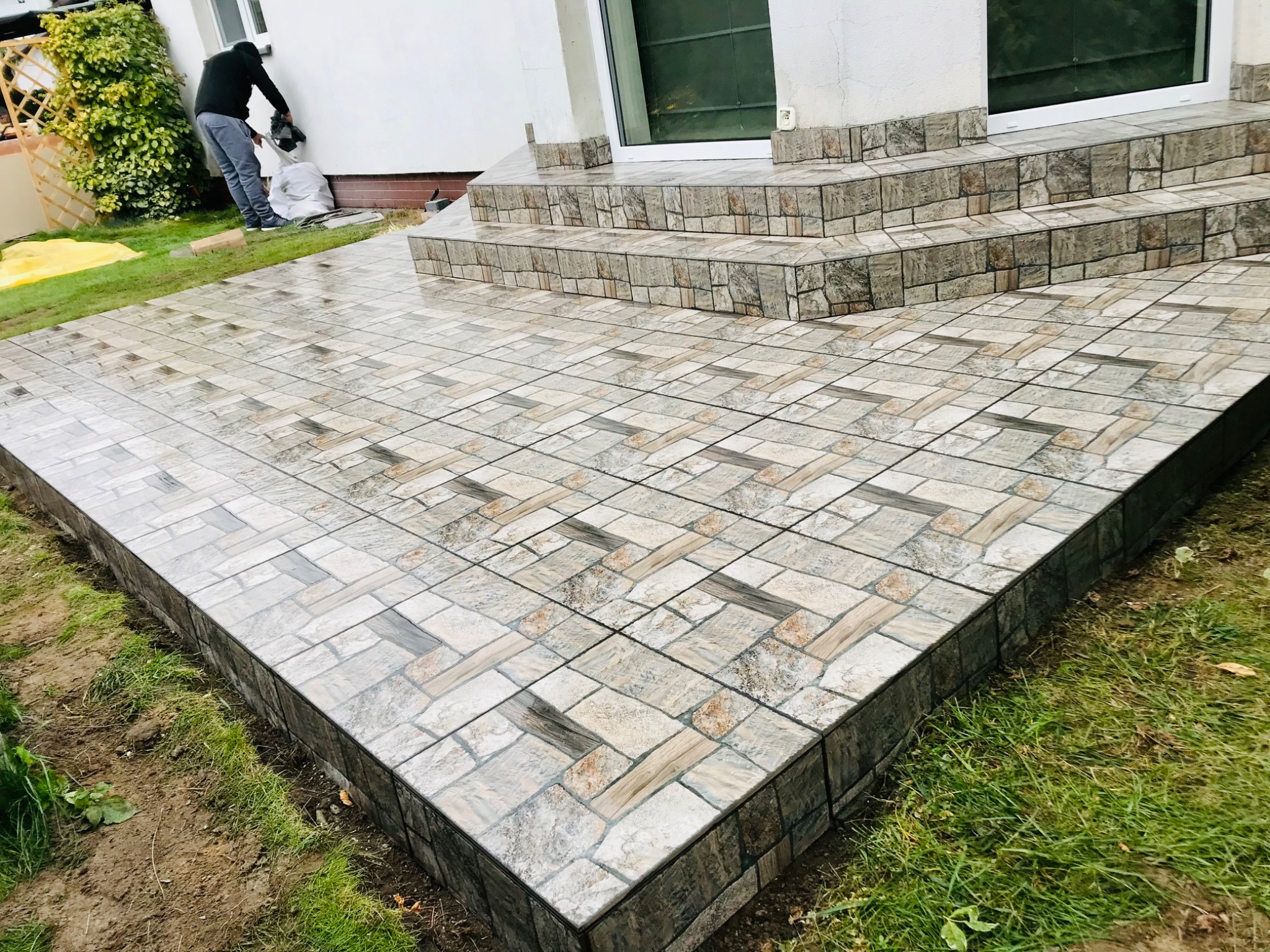 Wyłożony płytkami taras z niskimi schodami wejściowymi, widoczna osoba sprzątająca po pracy.