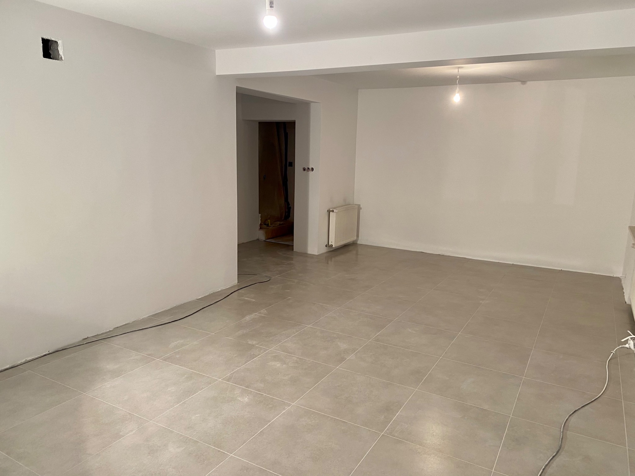 Jasne, przestronne wnętrze po zakończonych pracach, z beżową, kwadratową podłogą z płytek, białymi ścianami i widocznym grzejnikiem. W ścianie widoczny otwór instalacyjny.