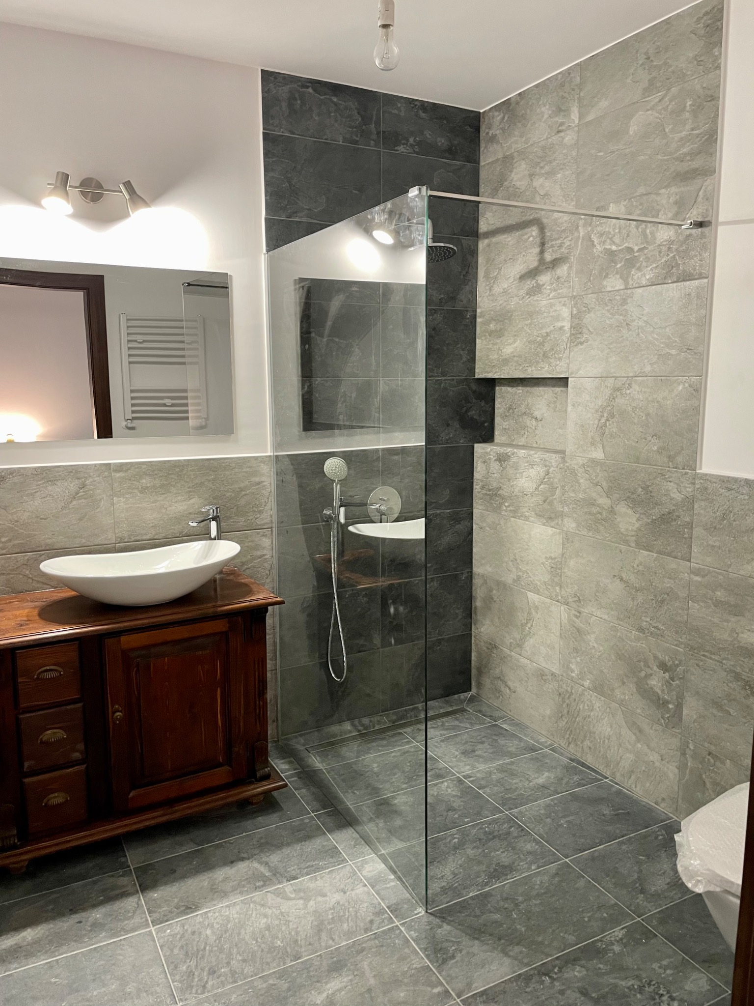 Wykończona łazienka z kabiną prysznicową typu walk-in, umywalką nablatową na drewnianej szafce w stylu retro, szare płytki imitujące kamień na ścianach i podłodze, oraz minimalistycznym oświetleniem.
