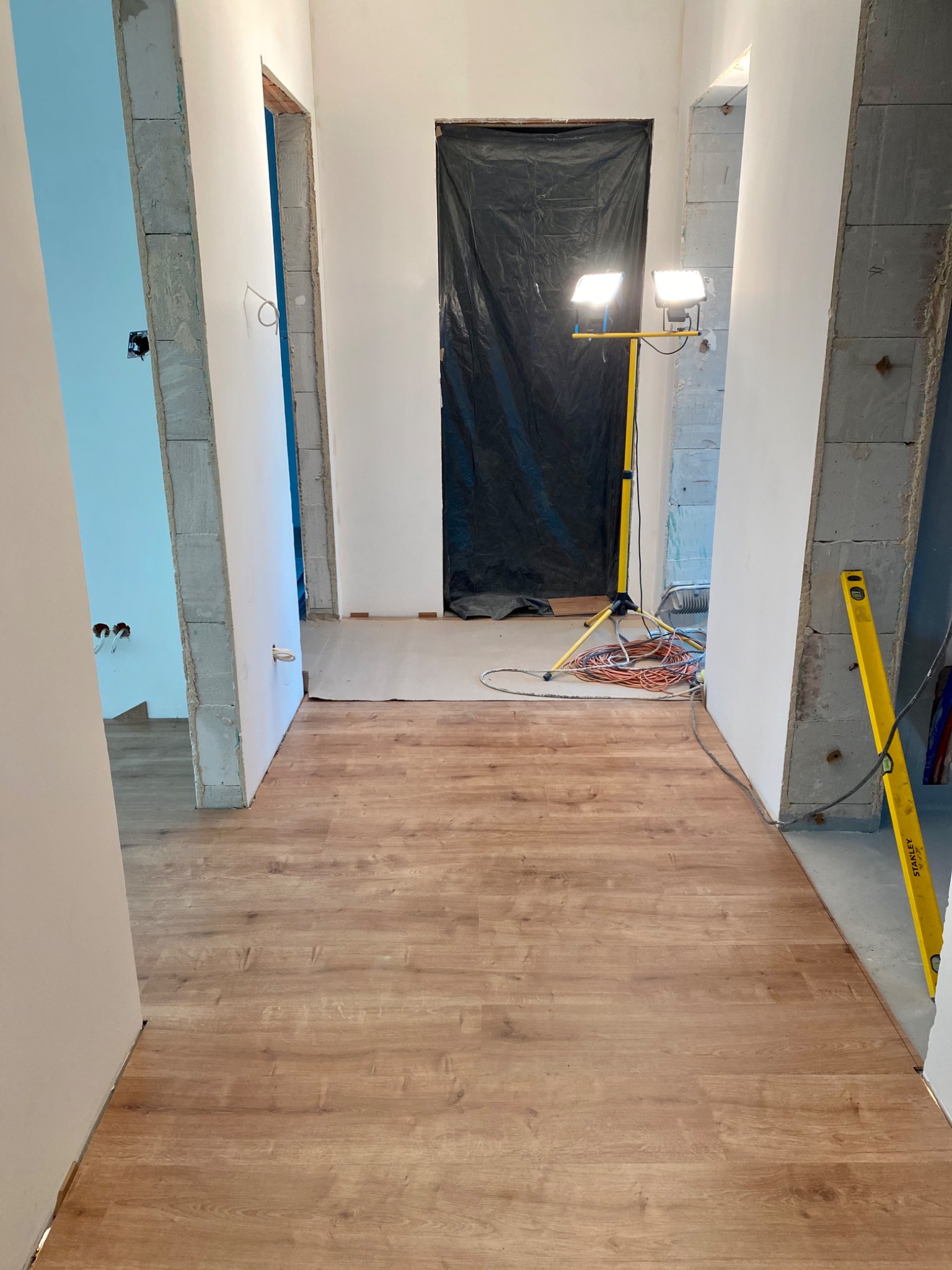 Korytarz w trakcie prac wykończeniowych: częściowo ułożona podłoga z paneli, widoczna poziomica Stanley na ścianie, lampa budowlana i kable, ściany w surowym stanie.