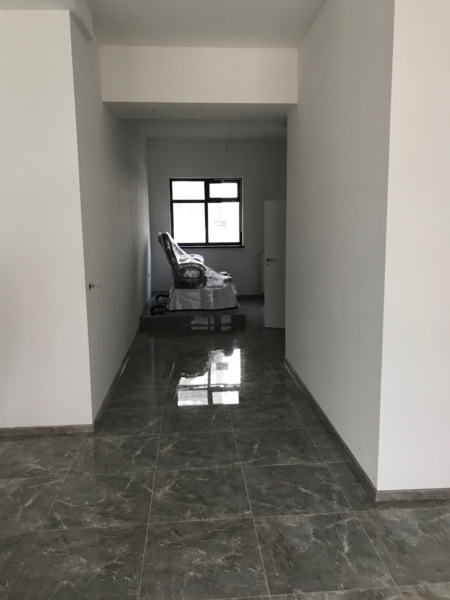 Wnętrze w trakcie prac wykończeniowych: szare marmurowe płytki podłogowe, białe ściany, okno z widokiem na zewnątrz, mebel zabezpieczony folią, widoczne niedokończone gniazdko elektryczne.