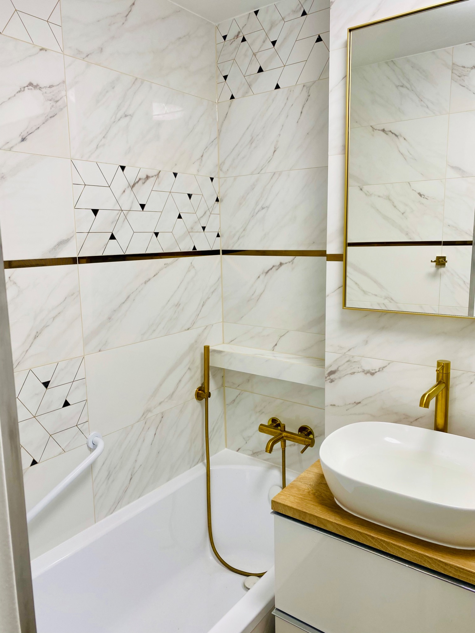 Biała łazienka z marmurowymi płytkami, akcentami w kolorze złota i geometrycznym wzorem na ścianie, obejmująca wannę z uchwytem, umywalkę nablatową i lustro w złotej ramie.