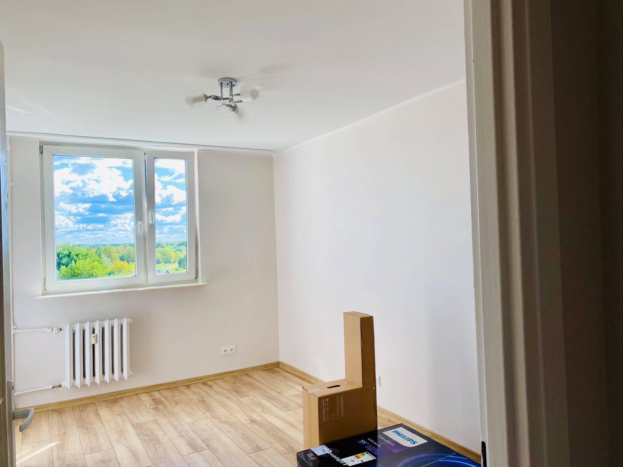 Pusty pokój z jasnymi ścianami, panelami podłogowymi w odcieniach drewna, białym grzejnikiem pod oknem z widokiem na zieleń i niebo, z kartonami po elektronice w rogu, sugerujący świeżo wykończone...