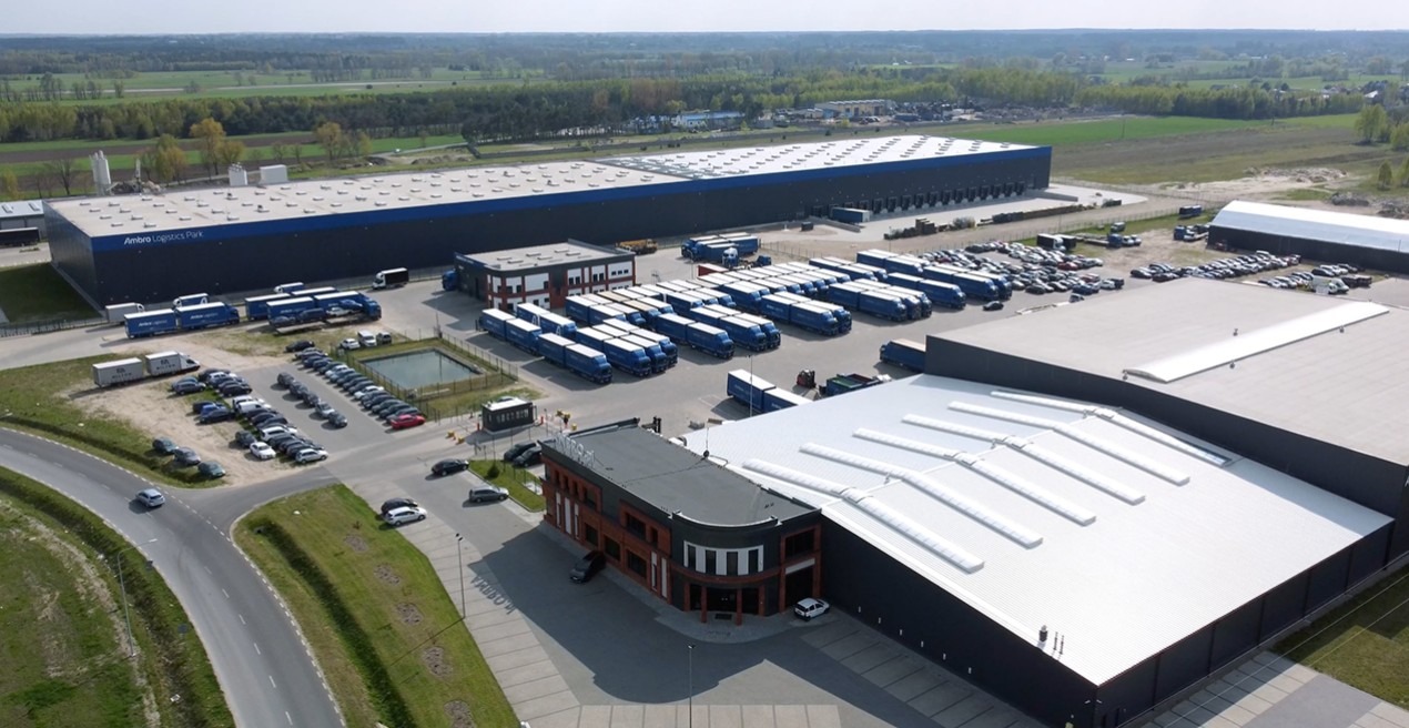 Szeroki widok z lotu ptaka na park logistyczny z wieloma niebieskimi ciężarówkami z naczepami, samochodami osobowymi i budynkami magazynowymi w słoneczny dzień.