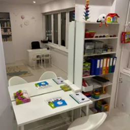 Gabinet logopedyczny: biały stół z materiałami edukacyjnymi, krzesła na kółkach, regał z zabawkami i pomocami dydaktycznymi, lustro na ścianie, w tle biurko i dywan z motywem chmur.