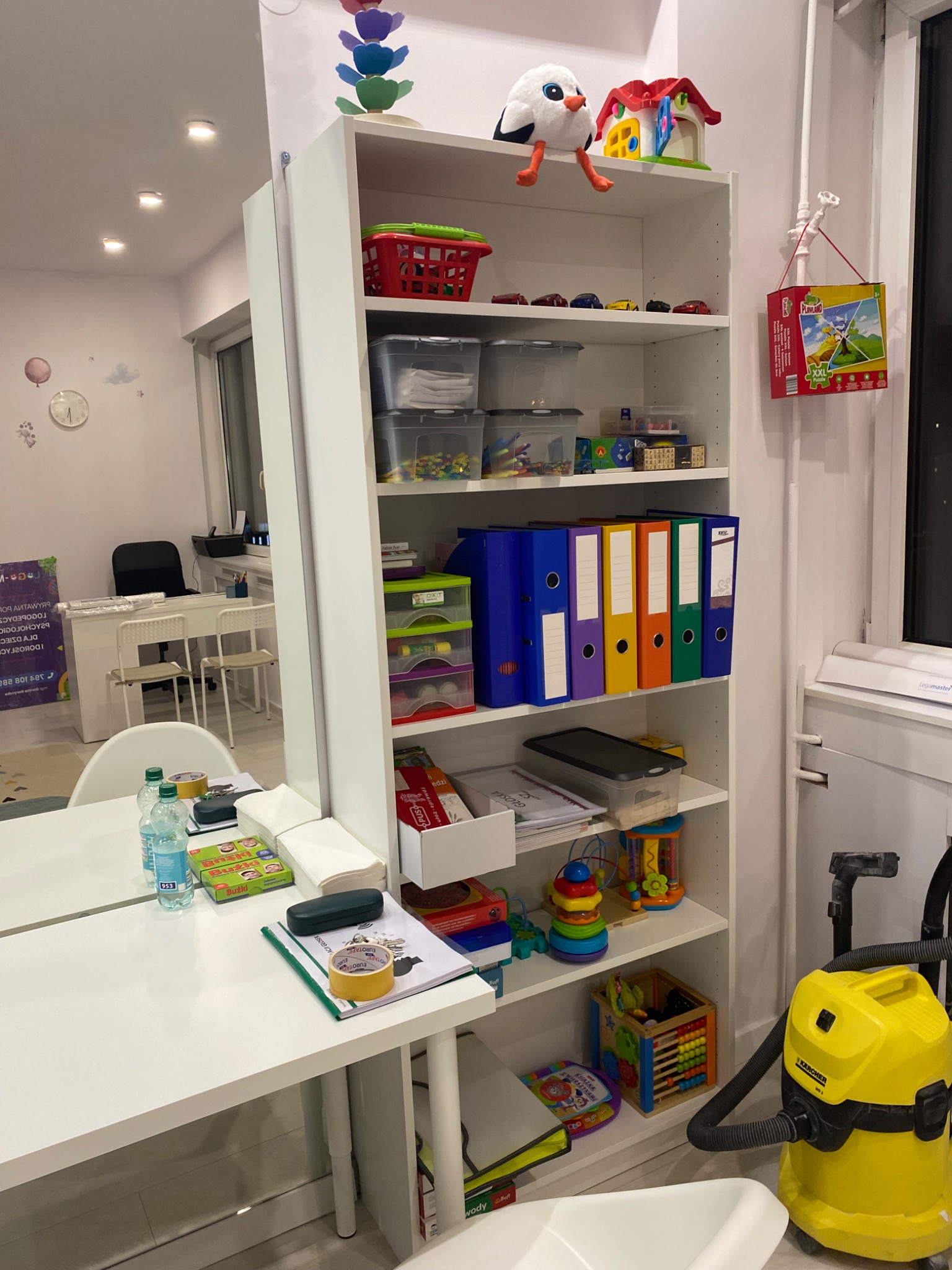 Gabinet terapeutyczny w Radomiu: białe biurko z materiałami edukacyjnymi, regał z zabawkami i pomocami dydaktycznymi, w tle lustro odbijające część pomieszczenia.