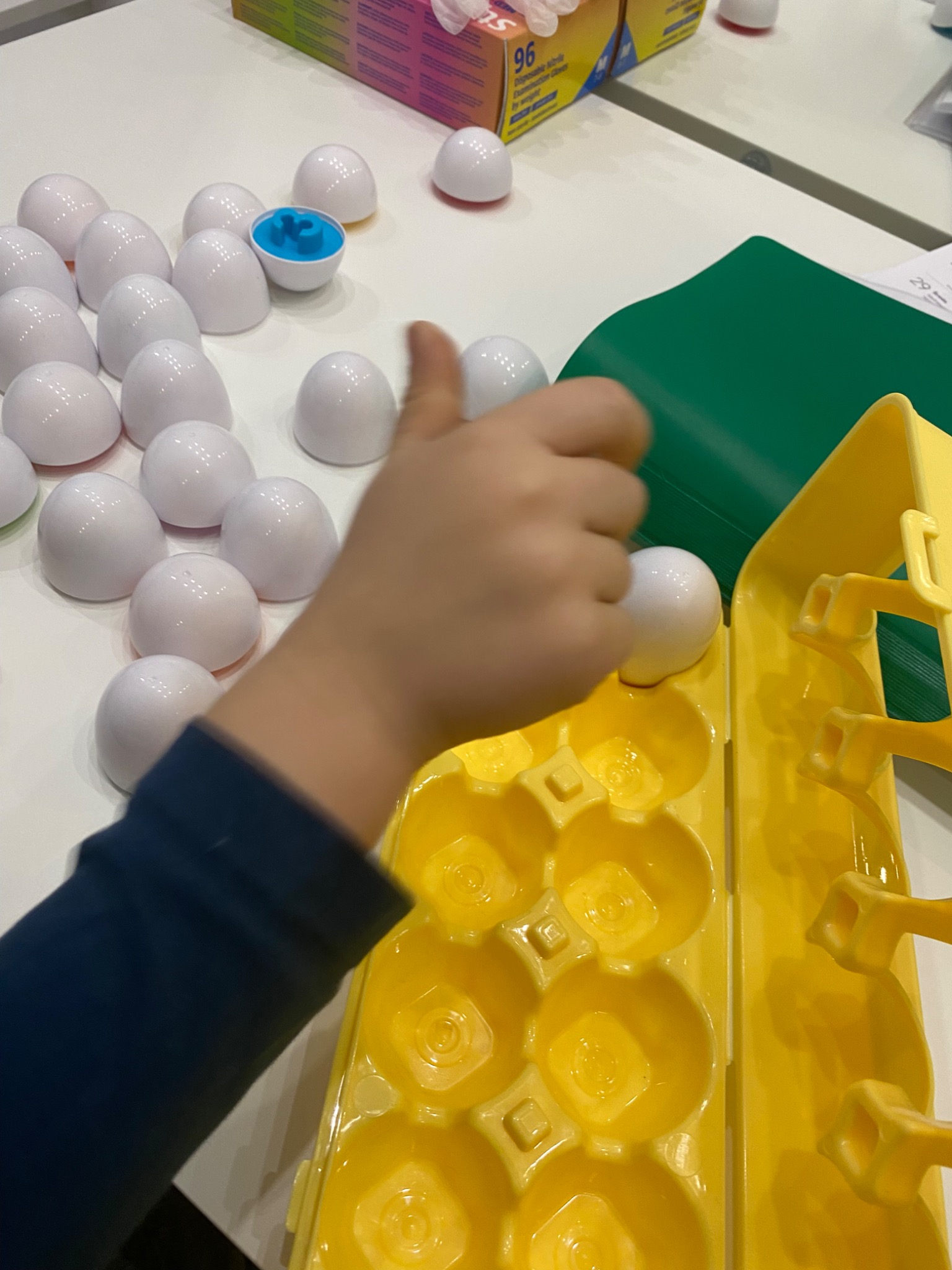 Dziecko umieszcza białą półkulę w żółtym pojemniku z wgłębieniami, obok leżą inne półkule, jedna z niebieskim elementem w środku, na białym stole.