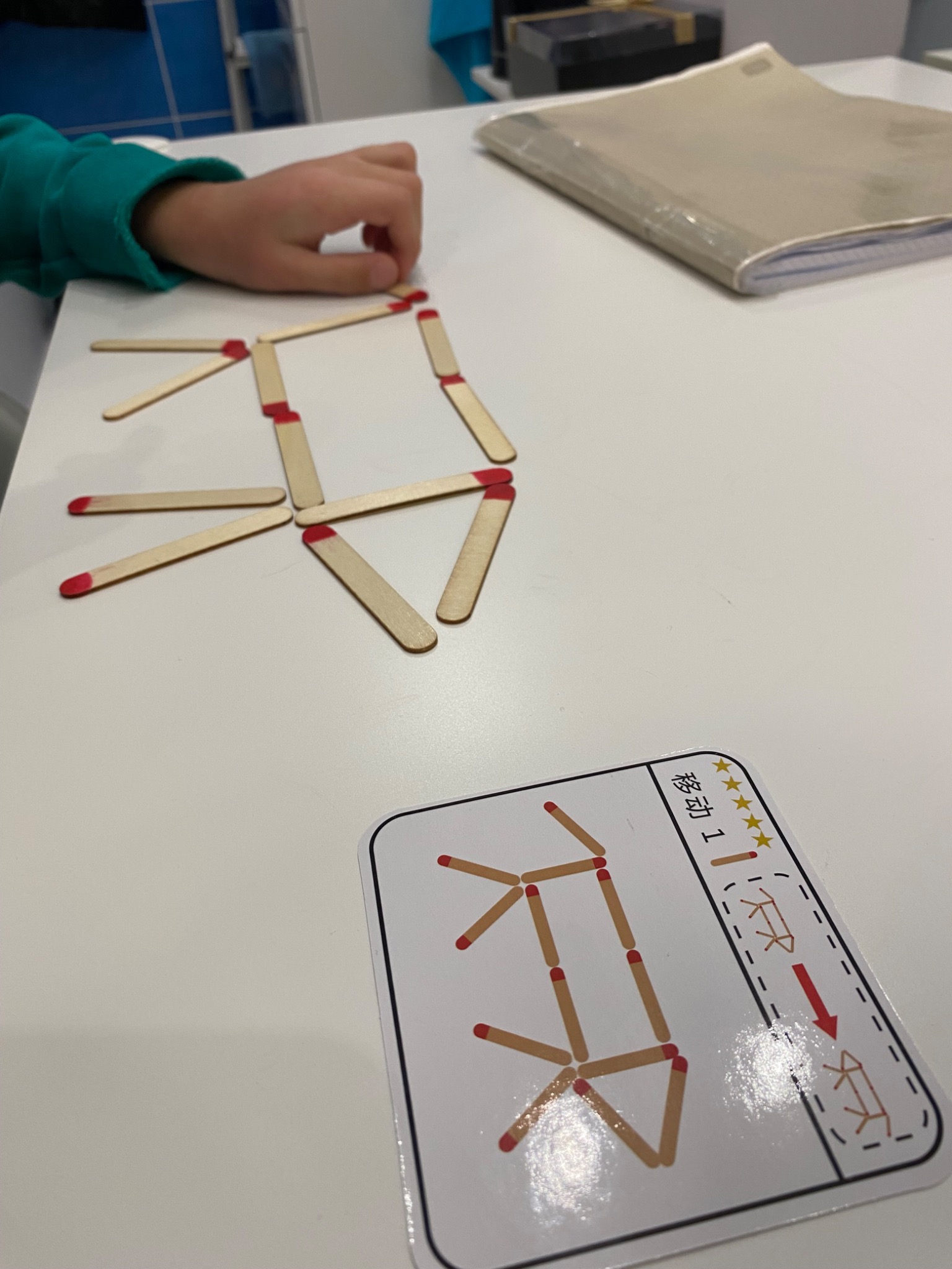 Dziecko układa figurę z patyczków z czerwonymi końcówkami, wzorując się na karcie z zadaniem logicznym na białym stole.