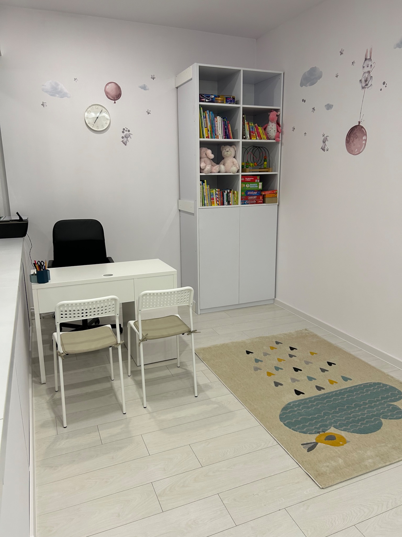 Jasny gabinet z biurkiem, krzesłami i regałem z książkami i zabawkami, dekorowany naklejkami ściennymi z motywem chmur i balonów.
