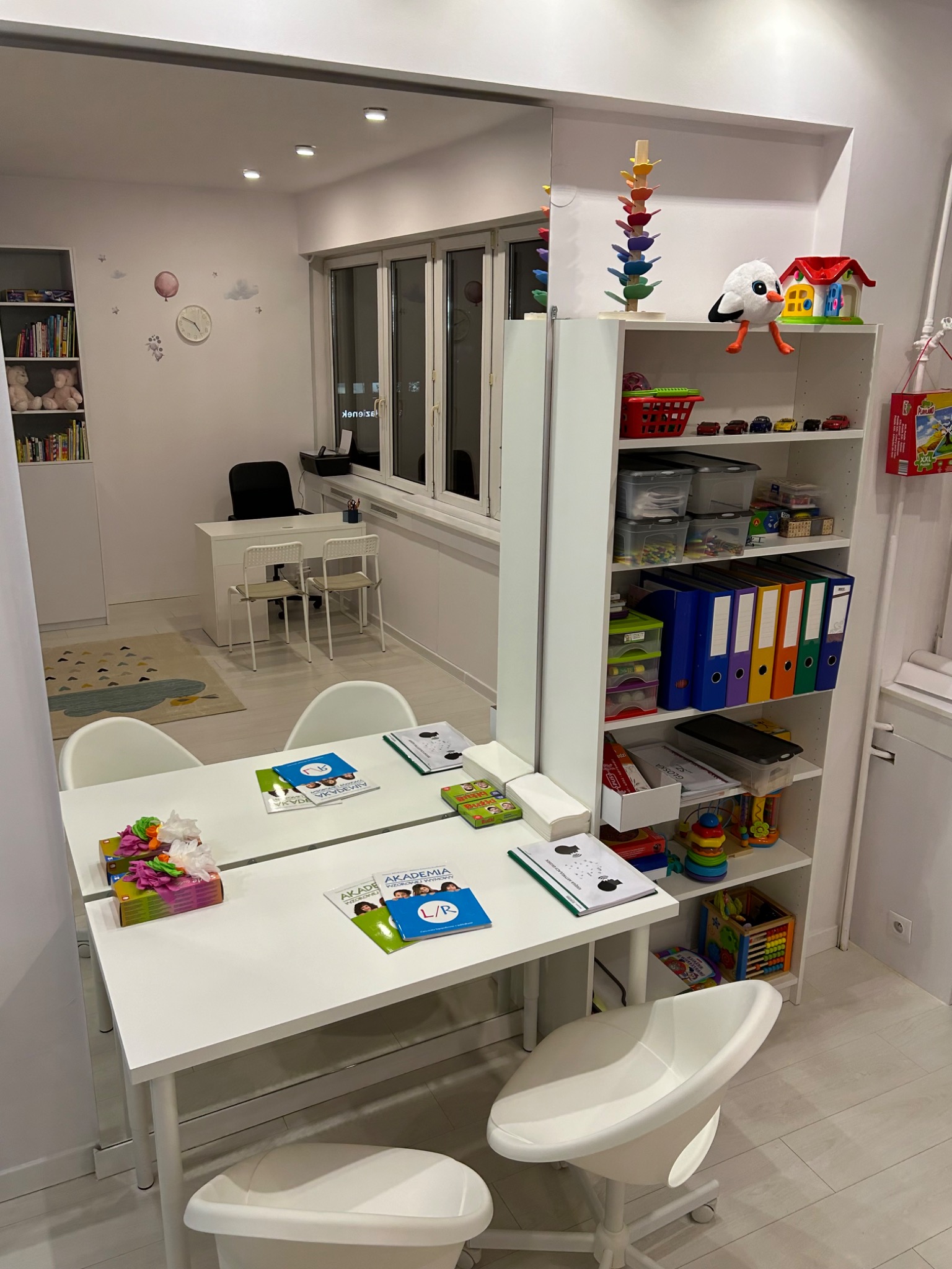 Gabinet logopedyczny: biały stół z materiałami edukacyjnymi, krzesła na kółkach, regał z zabawkami i pomocami dydaktycznymi, lustro na ścianie, w tle biurko i dywan z motywem chmur.
