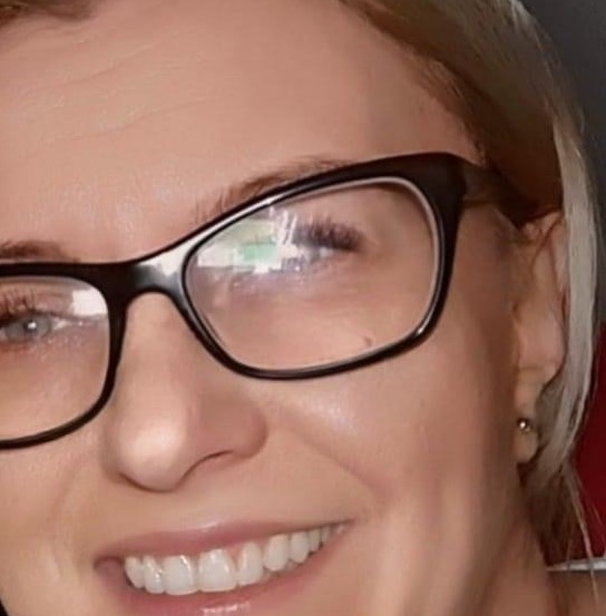 Portret kobiety w okularach z czarnymi oprawkami, uśmiechającej się i patrzącej w górę, widoczne odbicie w szkłach okularów.