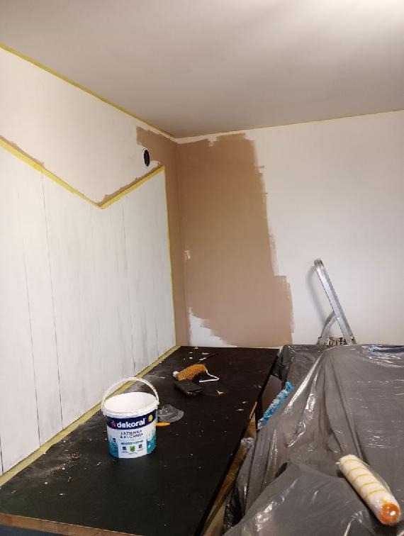 Fragment ściany w trakcie prac wykończeniowych: częściowo pomalowana na beżowo, zabezpieczona taśmą malarską przy suficie, widoczny kubełek z farbą Dekoral Łazienka & Kuchnia, wałek malarski...