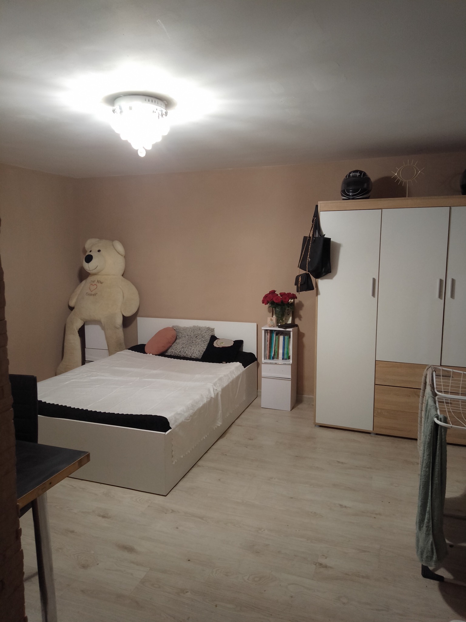 Pokój z łóżkiem, pluszowym misiem opartym o ścianę i szafą z kaskiem motocyklowym na górze, ściany pomalowane na beżowo.