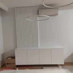 Biała szafka z czterema drzwiczkami pod klimatyzatorem, z dekoracyjną ścianką z pionowych listew. Na podłodze karton i folie ochronne, sugerujące świeży montaż.