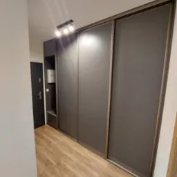 Wnętrze przedpokoju z dużą, przesuwną szafą wnękową o matowych, grafitowych frontach i drewnianych obramowaniach, oświetloną trzema punktowymi lampami sufitowymi, z widocznym fragmentem zabudowy...