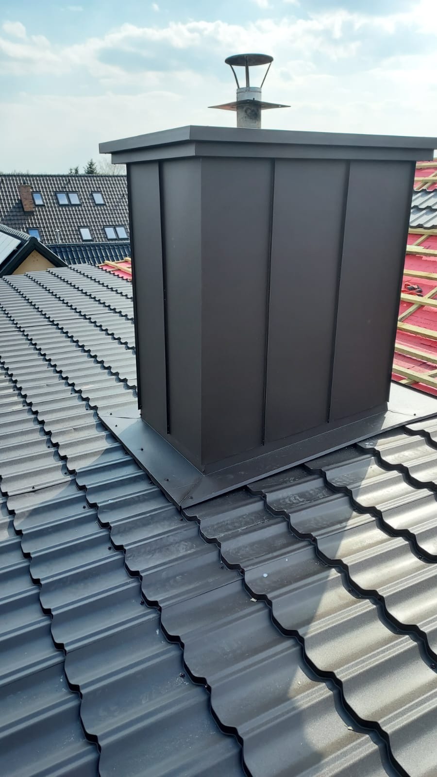 Nowoczesny, grafitowy komin obłożony blachą na dachu z blachodachówki w kolorze grafitowym, widoczny fragment dachu sąsiedniego budynku z czerwoną membraną.