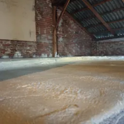 Poddasze z widoczną warstwą natryskowej piany izolacyjnej na podłodze, ceglana ściana z fragmentami starego tynku, drewniane elementy konstrukcji dachu.
