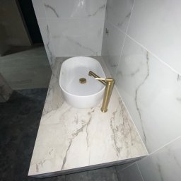 TILER Oleksandr Lebediev - Biała umywalka nablatowa z baterią w kolorze złota, na blacie imitującym marmur. W tle białe płytki z szarymi żyłkami i fragment czarnej podłogi.