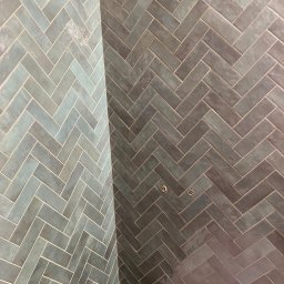 TILER Oleksandr Lebediev - Ściana wyłożona błyszczącymi, podłużnymi płytkami w odcieniach szarości i turkusu, ułożonymi we wzór jodełki; widoczne dwa otwory na instalacje.