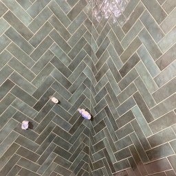 TILER Oleksandr Lebediev - Ściana w łazience wyłożona błyszczącymi, zielono-szarymi płytkami ułożonymi w jodełkę. Widoczne wystające rury z zaślepkami.