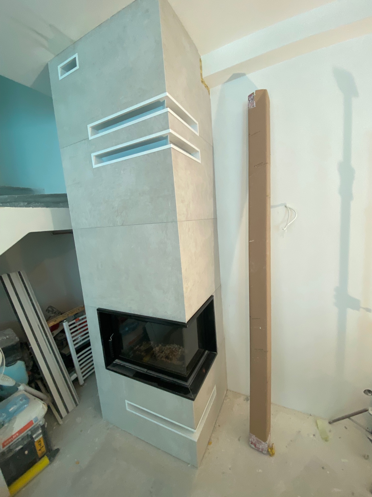 Nowoczesny kominek obłożony płytami w odcieniach szarości, z dekoracyjnymi wnękami i czarnym wkładem, w trakcie wykańczania. Widoczne materiały budowlane.