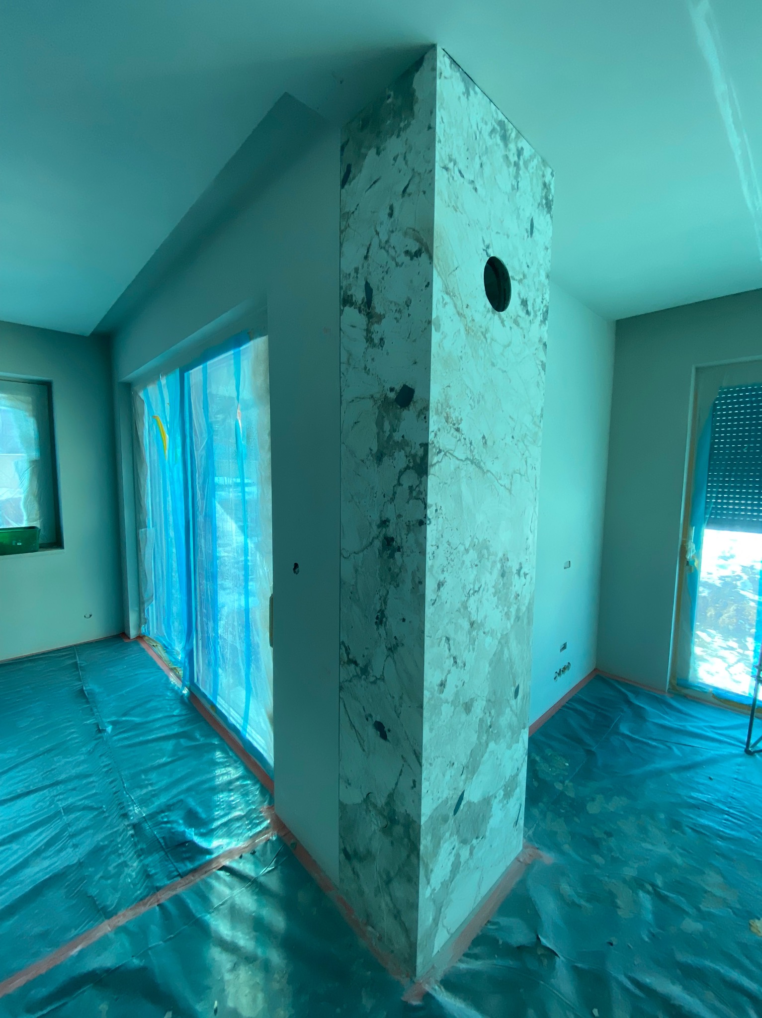 Wykończony filar z marmurowym wzorem w pokoju, zabezpieczona folią podłoga i okna. W ścianie okrągły otwór. Prace wykończeniowe w trakcie.