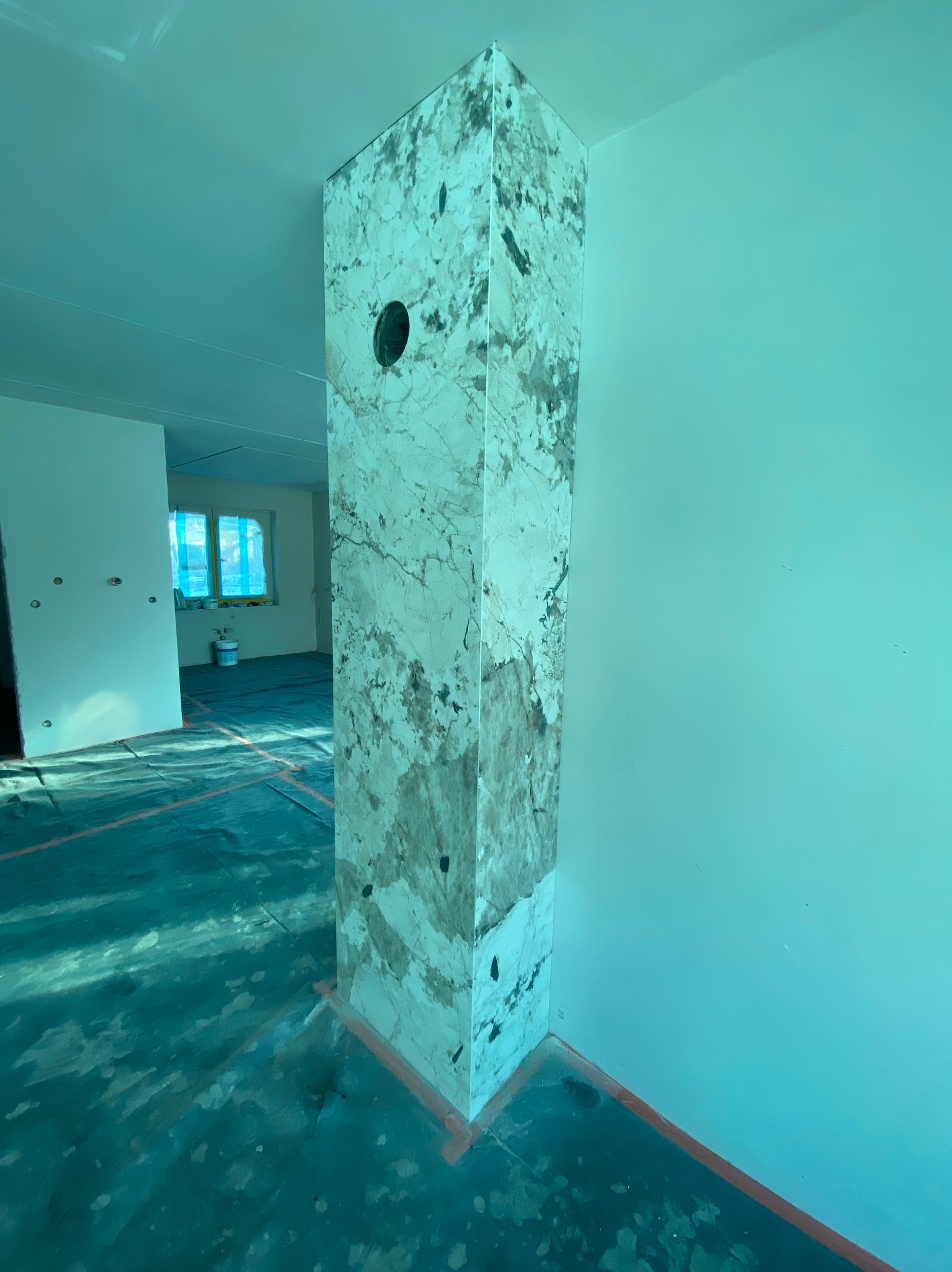 Wykończony filar z efektem marmuru w nowym wnętrzu. Widoczne zabezpieczenia podłogi i surowy stan ścian. Otwór w filarze sugeruje instalację.