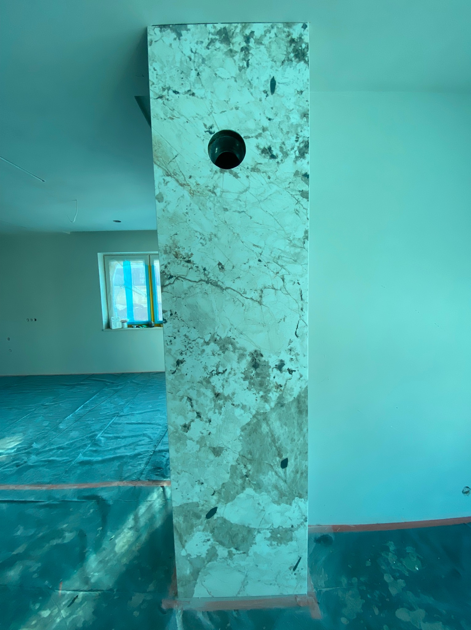 Kolumna z efektem marmuru z otworem na rurę w remoncie, zabezpieczona folią podłoga, okno w tle. Nietypowe wykończenie ściany z elementami instalacji.