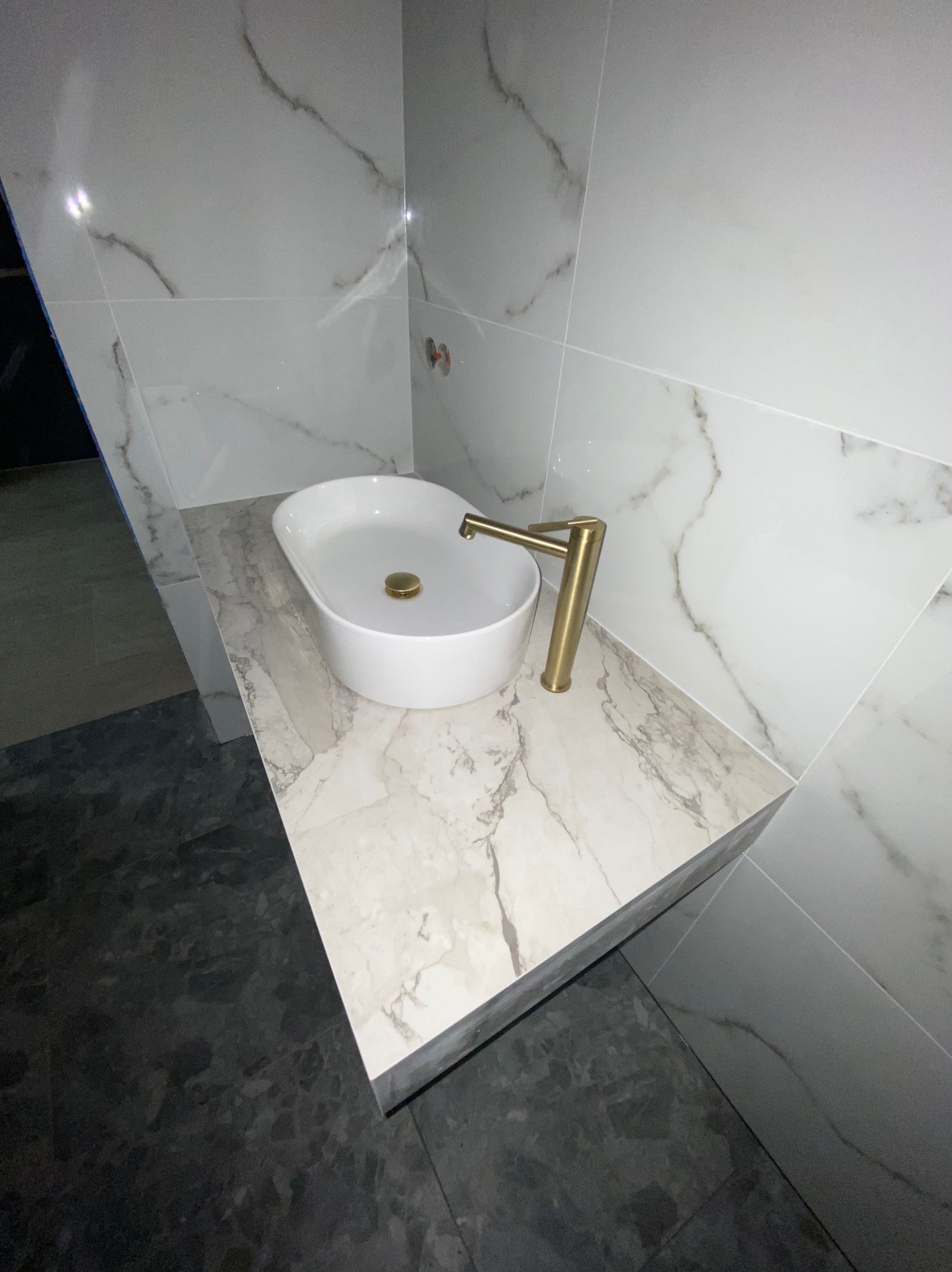 Nowoczesna łazienka z białą umywalką nablatową, złotą armaturą i marmurowymi płytkami na ścianach i blacie. Ciemna podłoga podkreśla kontrast.