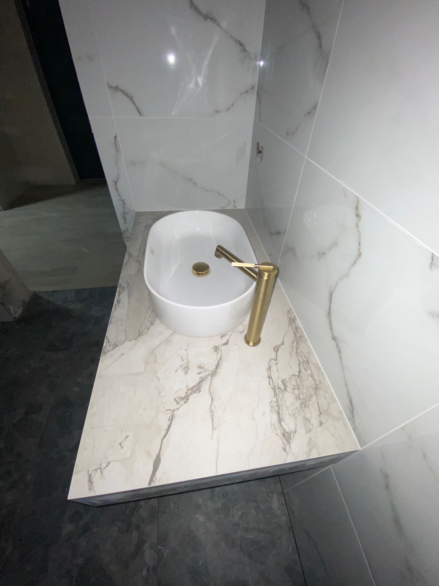 Biała umywalka nablatowa z baterią w kolorze złota, na blacie imitującym marmur. W tle białe płytki z szarymi żyłkami i fragment czarnej podłogi.