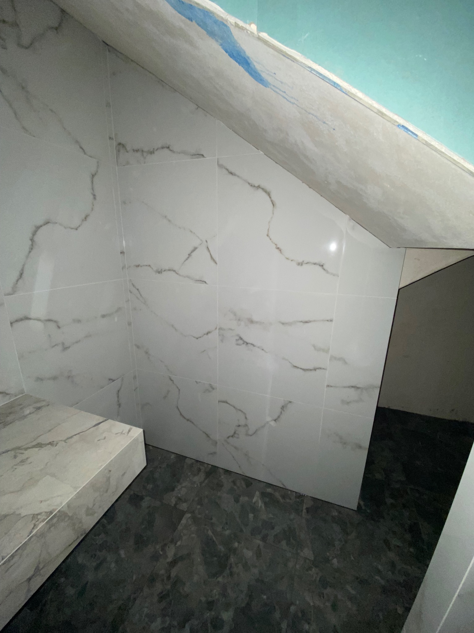 Wykończona łazienka na poddaszu: białe płytki marmurowe na ścianach skośnych i szare płytki podłogowe. Widoczne łączenia płytek i surowy stan sufitu.