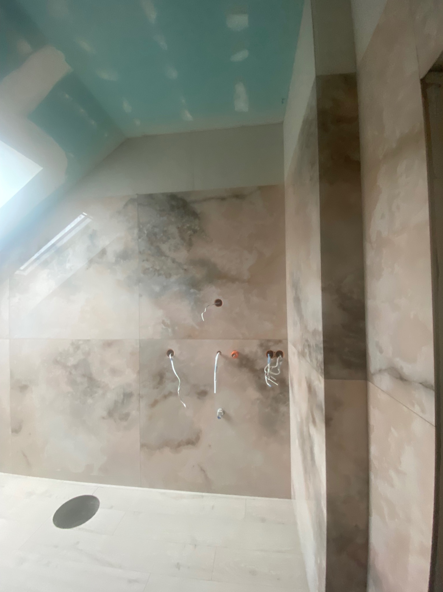 Łazienka w trakcie remontu: marmurowe płytki na ścianach, sufit z płyt karton-gips, widoczne instalacje wodne i odpływ podłogowy. Etap prac glazurniczych i wykończeniowych.