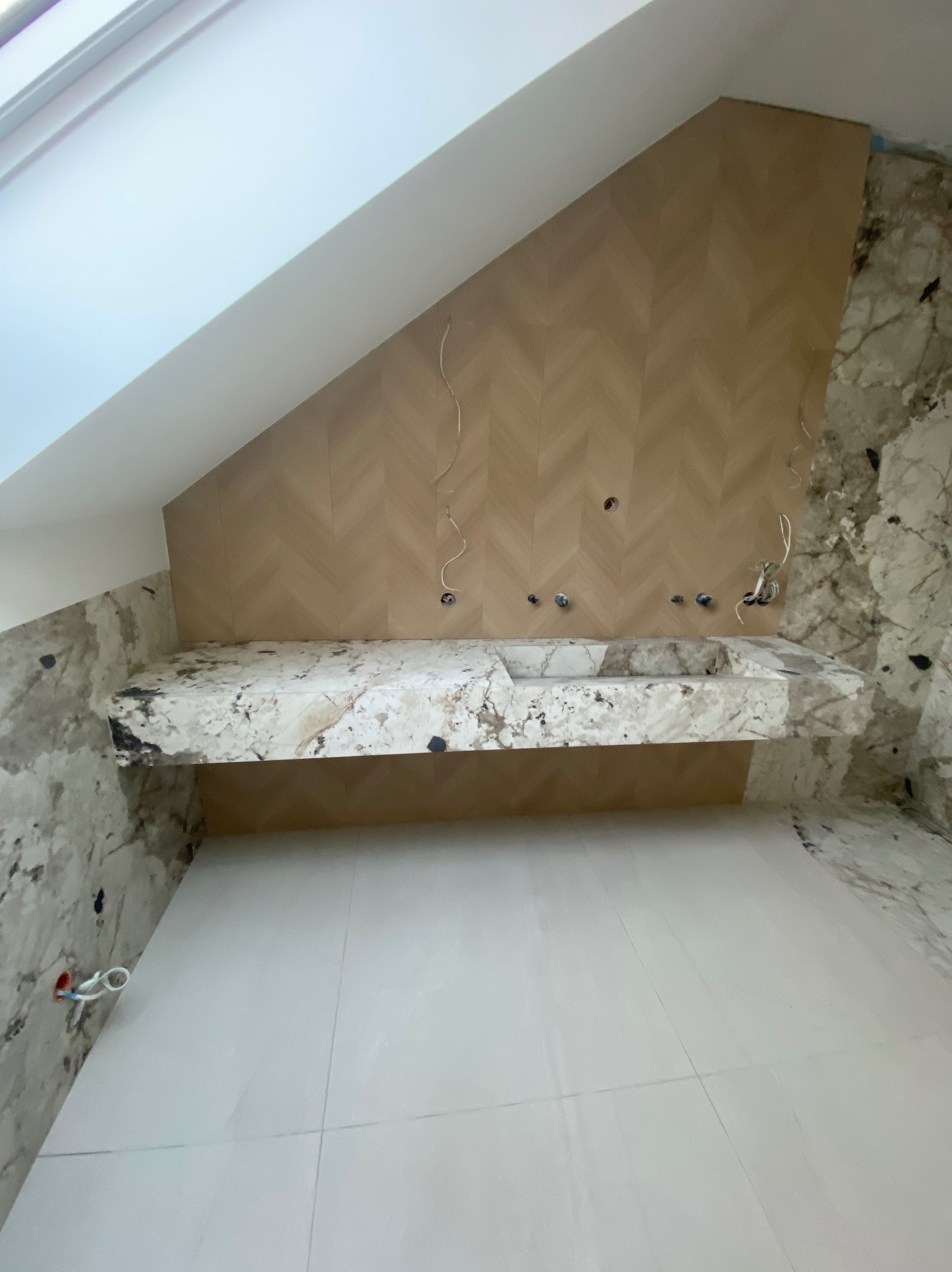 Łazienka w trakcie wykończenia: marmurowe ściany, drewniana mozaika na skośnej ścianie poddasza, blat umywalki z marmuru. Widoczne przewody instalacji.