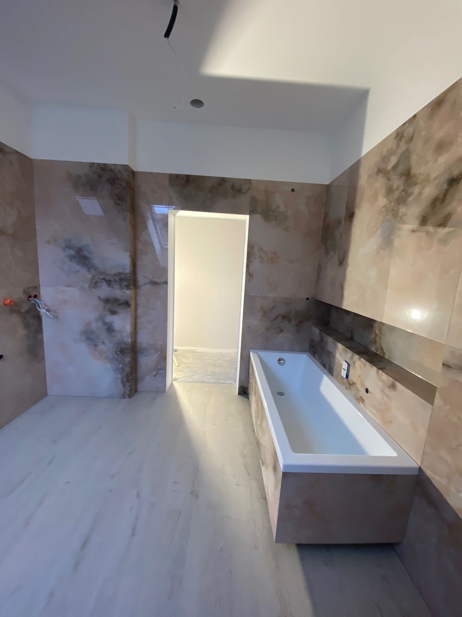 Wykończona łazienka z wanną w zabudowie i marmurkowymi płytkami na ścianach. Widoczne podłączenia elektryczne i fragment otwartych drzwi do kolejnego pomieszczenia.