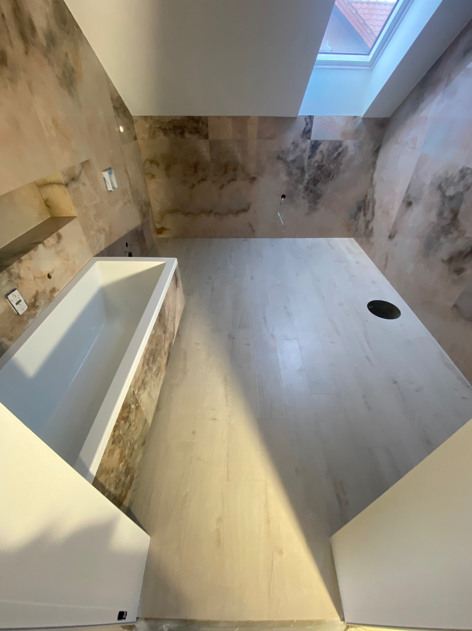 Wykończona łazienka na poddaszu z beżowymi płytkami imitującymi marmur, białą wanną i oknem dachowym. Widok z góry.