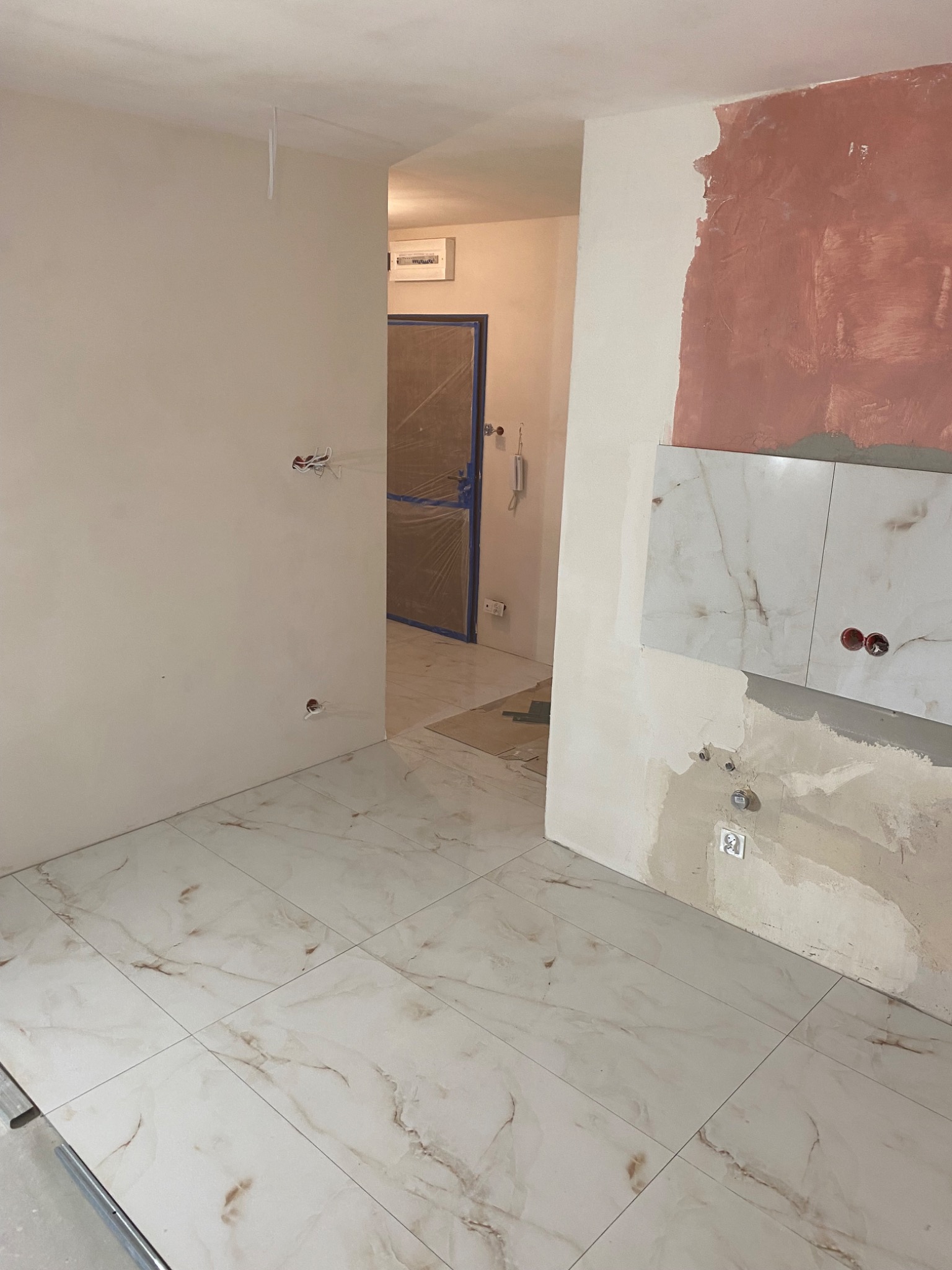 Wnętrze w trakcie prac wykończeniowych: białe płytki podłogowe z marmurkowym wzorem, częściowo wykończona ściana z widocznymi instalacjami elektrycznymi i fragmentem ściany pomalowanym na różowo...