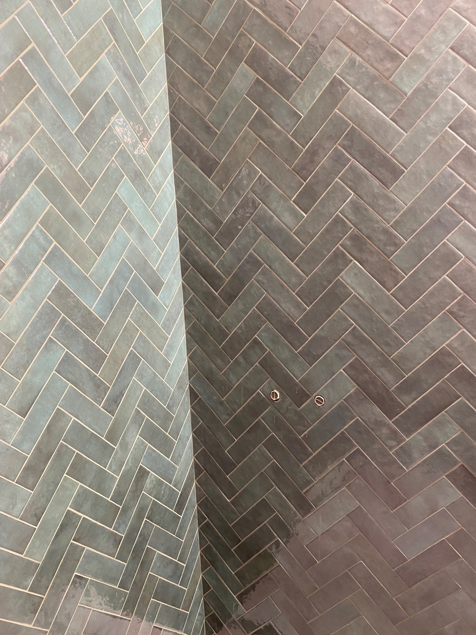 Ściana wyłożona błyszczącymi, podłużnymi płytkami w odcieniach szarości i turkusu, ułożonymi we wzór jodełki; widoczne dwa otwory na instalacje.