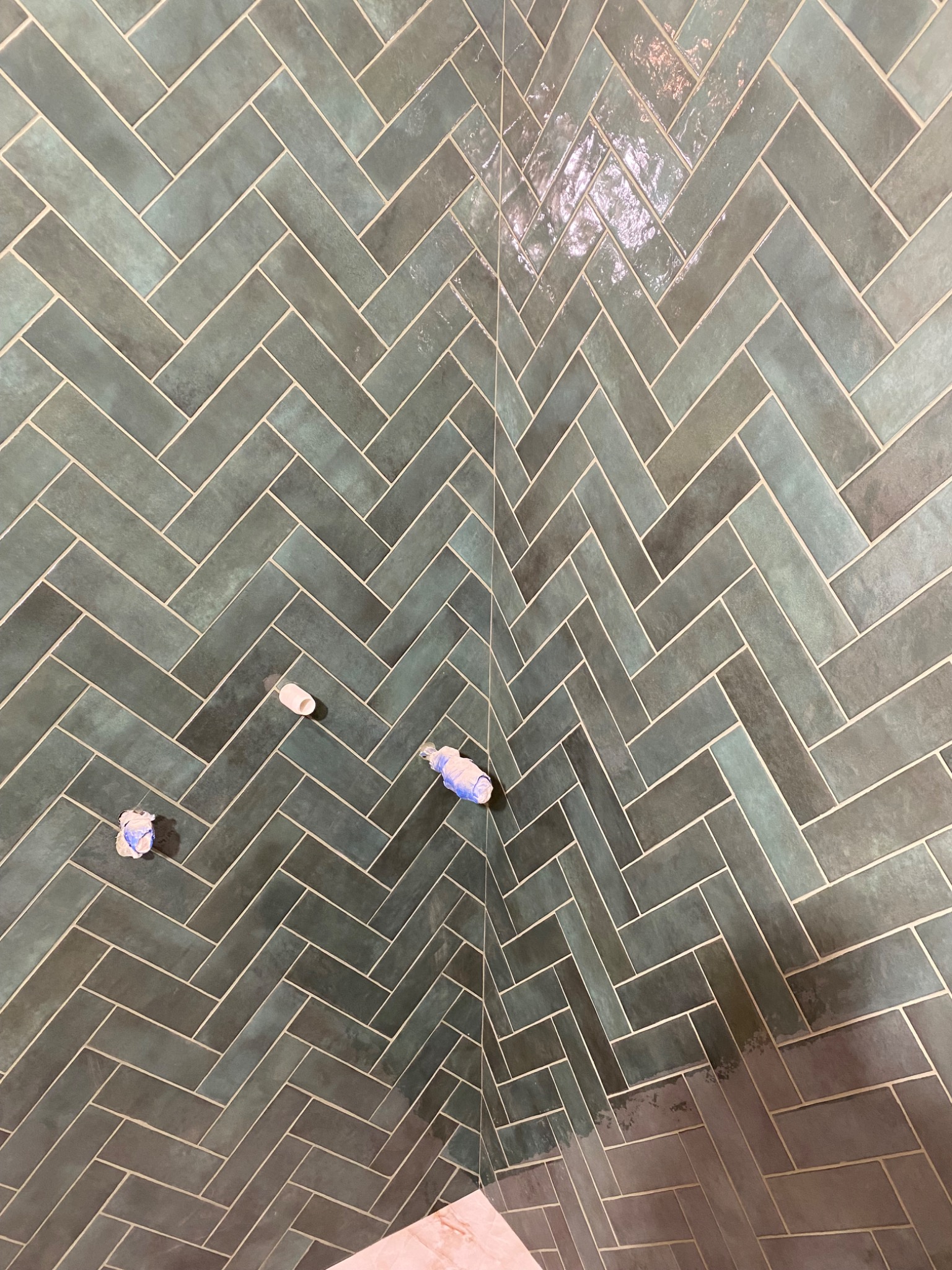 Ściana w łazience wyłożona błyszczącymi, zielono-szarymi płytkami ułożonymi w jodełkę. Widoczne wystające rury z zaślepkami.