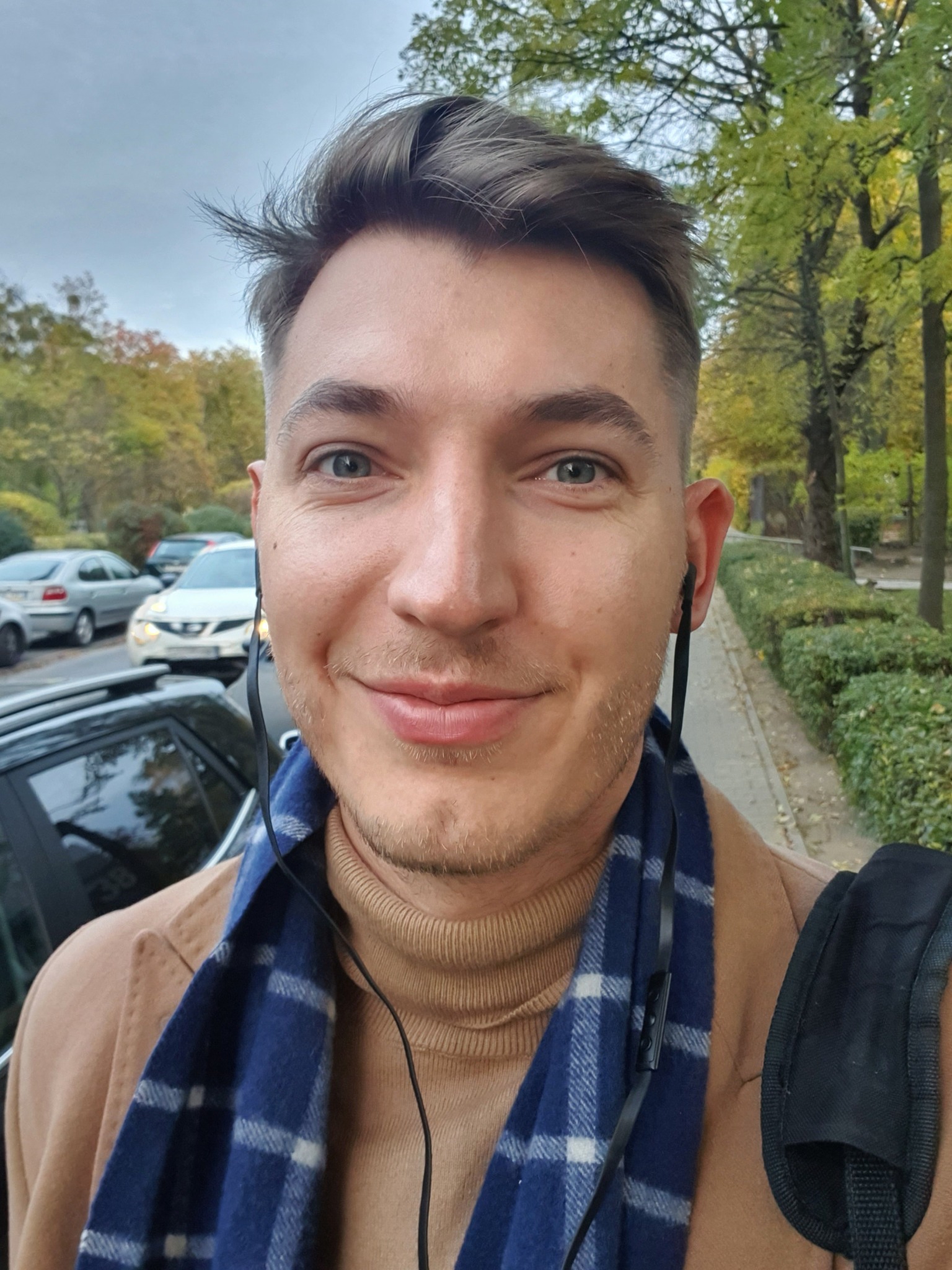 Portret uśmiechniętego mężczyzny w jesiennej scenerii miejskiej, w beżowym płaszczu, golfie i niebieskim szaliku w kratę, z słuchawkami w uszach, na tle ulicy z samochodami i zielenią.