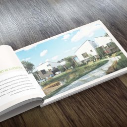 A2 studio - Otwarta broszura reklamowa leżąca na drewnianej powierzchni, prezentująca wizualizację nowoczesnego osiedla domów jednorodzinnych z zielenią i jeziorkiem.